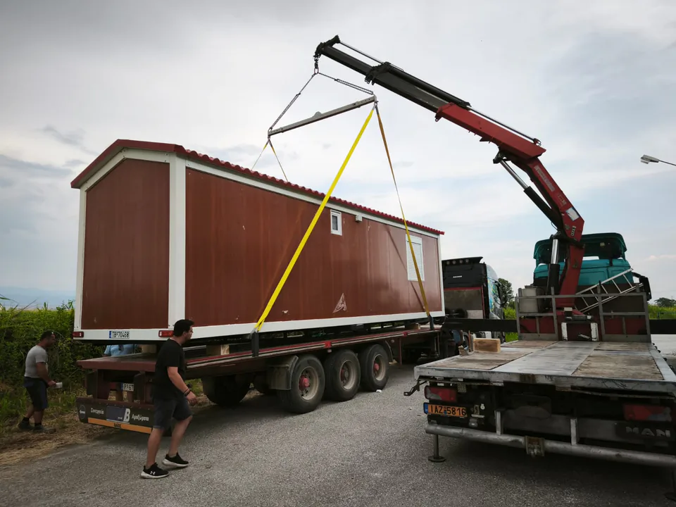 Crane moving a long mobile home between trucks