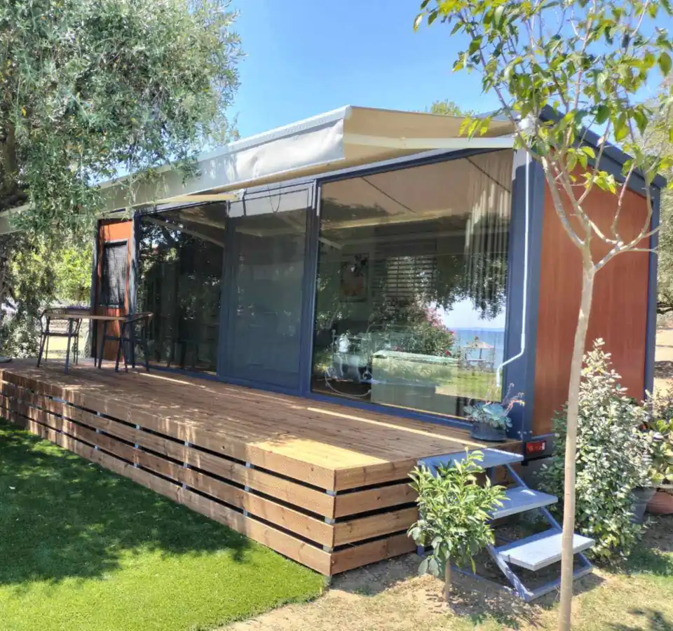 Nature Line mobile home with glass walls, an awning and a timber deck under olive trees, with a sea view
