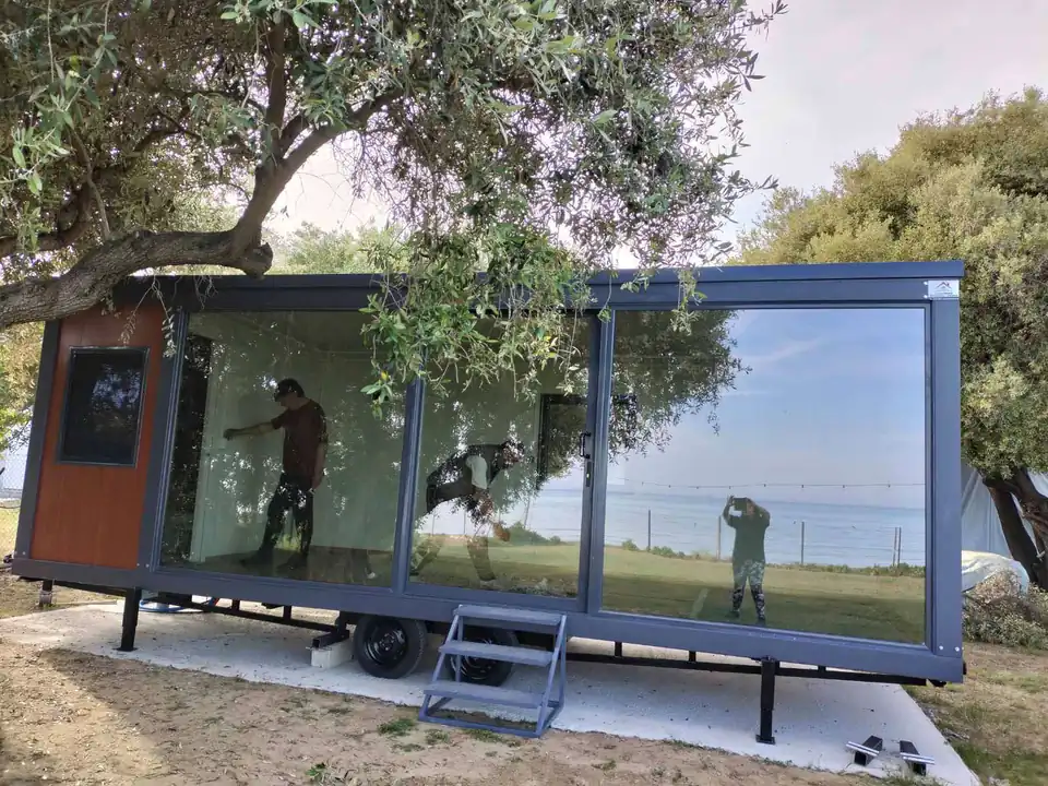 Glass-walled mobile home under an olive tree, with a sea view